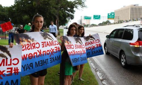 年轻女孩在路边举着标语，上面写着“617888九五至尊娱乐危机，#埃克森知道让他们付出代价。."